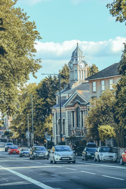 A street scene in Hornchurch featuring a historic building with a clock tower and a decorative spire, surrounded by leafy trees and residential buildings. Parked cars line the roadside, and the sky is clear with a few clouds. Inside the property, there are visible cardboard boxes, protected furniture wrapped in plastic and blankets, and a moving trolley nearby. The scene suggests an ongoing home relocation or furniture transport operation as part of a local house removal service, with the loading process possibly taking place near the pavement or driveway. A man with van company, Man with Van Hornchurch, appears to be involved in the packing and moving activities, with equipment such as straps and trolleys supporting the operation within the context of house removals in the Hornchurch RM11 area.