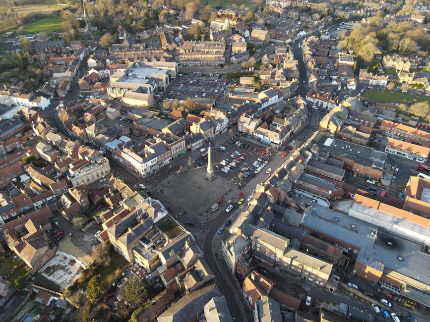 An aerial view of a town centre showing a large, open town square with a tall monument or column in the middle, surrounded by parked cars and a mix of historic and modern buildings. Narrow streets radiate from the square, lined with shops, houses, and commercial properties. Residential areas with terraced houses extend outward, with some rooftops covered in red tiles and others in darker materials. Trees and greenery are visible along the streets and in the distance, providing a scenic backdrop. The lighting suggests a bright day with clear skies, suitable for home relocation and furniture transport activities. Man with Van Hornchurch typically conducts home removals in such urban environments, carefully managing the loading process of furniture and packed boxes onto vans, including the use of trolleys and protective blankets for safe transport during house removals in Hornchurch.