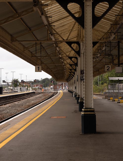 Empty train platform at Hornchurch station with a covered canopy supported by white columns, numbered signs for platform 1 and 2, yellow tactile paving along the edge, and railway tracks extending into the distance under an overcast sky. In the background, there are distant buildings and some greenery, while a small waiting area with benches is visible on the right side. The scene is relevant to house removals and moving logistics, illustrating the station environment associated with transportation services, such as those provided by Man with Van Hornchurch for home relocation and furniture transport.