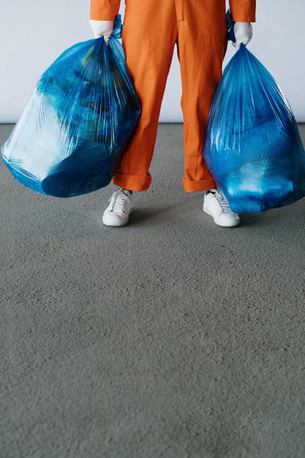 A male worker, wearing a high-visibility orange vest and dark pants, is standing at the rear of a large red garbage collection truck on a city street, during a daytime clean-up operation. The truck's 
