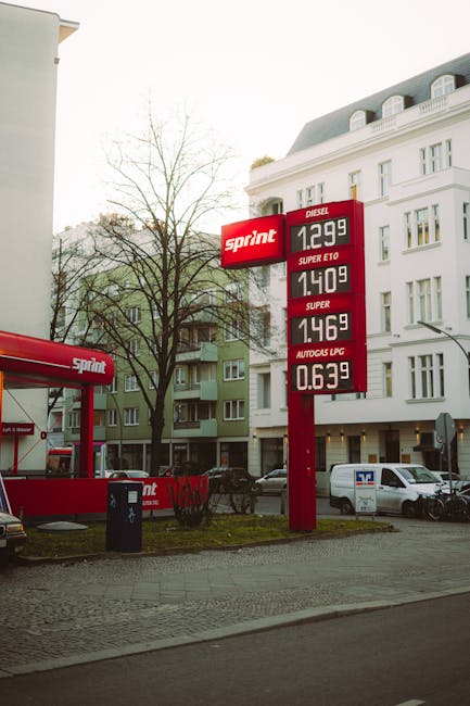 The image shows a roadside scene outside a residential building in Hornchurch, featuring a large red fuel station sign displaying prices for diesel, petrol, and LPG fuel types. Adjacent to the sign, a red man with a van branded vehicle is parked on a paved area next to a black trash bin. The van is part of a home relocation or furniture transport process associated with Man with Van Hornchurch, with the van's rear door possibly open for loading or unloading belongings. Behind the scene, modern multi-storey buildings with white and light green facades and multiple balconies are visible, along with a leafless tree indicating a colder season or early spring. Several parked cars and a white delivery van are positioned along the street, providing context for urban moving activities. The scene is lit with natural daylight, emphasizing the practical environment of logistics involved in packing and moving services.