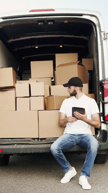 A man wearing a white t-shirt, blue jeans, white sneakers, and a black cap is sitting on the edge of an open van trunk during a home relocation process. Inside the van, there are numerous cardboard moving boxes of various sizes, some stacked on top of each other, and a few larger boxes placed on the floor of the vehicle. The boxes are sealed with packing tape, and some are wrapped in plastic for protection. The loading area appears to be outdoors on a paved surface, with the vehicle positioned near a building entrance, as indicated by the door visible at the back of the van. The man is holding a tablet or clipboard, possibly reviewing inventory or planning the next step in the furniture transport or packing and moving process. This scene illustrates the preparation phase of house removals, with the focus on the efficient organization and handling of packing materials as part of professional removals services by Man with Van Hornchurch.