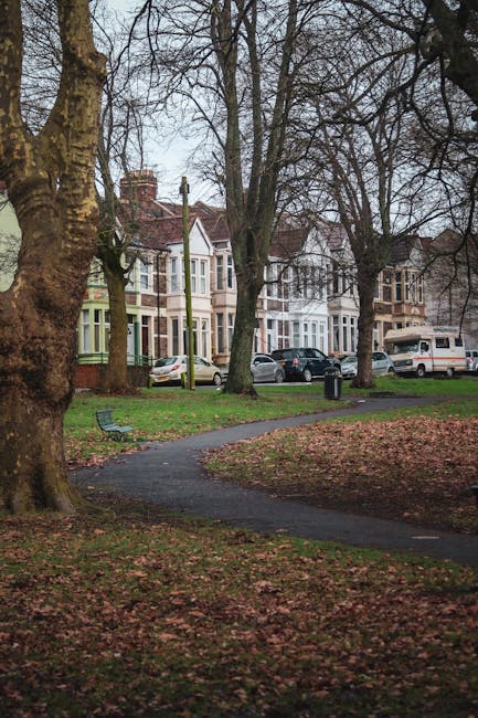 A residential street view showing a row of Victorian-style terraced houses with bay windows and brick exteriors, situated behind a small park with a paved footpath. The park features large, mature trees with bare branches, indicating late autumn or early winter, and fallen leaves scattered across the grass and pathway. Park benches and a trash bin are visible along the walkway, which curves through the green space. Several parked cars, including sedans and vans, are on the street in front of the houses. The scene is captured during daytime with overcast sky, and there is no visible activity or moving objects related to house removals or furniture transport. This image exemplifies a typical UK suburban neighborhood environment, suitable for a house removal or relocation service overview provided by Man with Van Hornchurch.