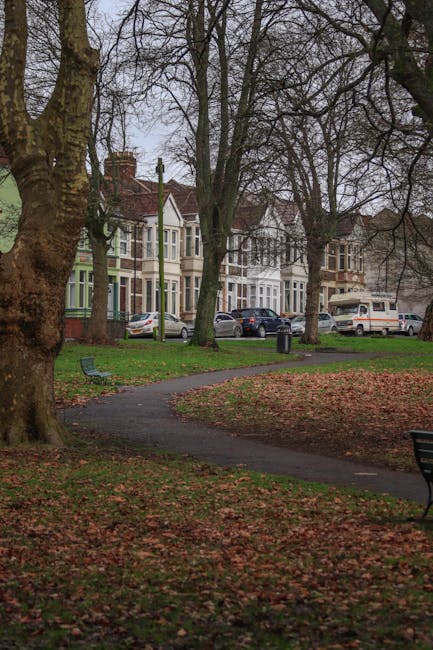 A residential street view showing a row of Victorian-style terraced houses with bay windows and brick exteriors, situated behind a small park with a paved footpath. The park features large, mature trees with bare branches, indicating late autumn or early winter, and fallen leaves scattered across the grass and pathway. Park benches and a trash bin are visible along the walkway, which curves through the green space. Several parked cars, including sedans and vans, are on the street in front of the houses. The scene is captured during daytime with overcast sky, and there is no visible activity or moving objects related to house removals or furniture transport. This image exemplifies a typical UK suburban neighborhood environment, suitable for a house removal or relocation service overview provided by Man with Van Hornchurch.