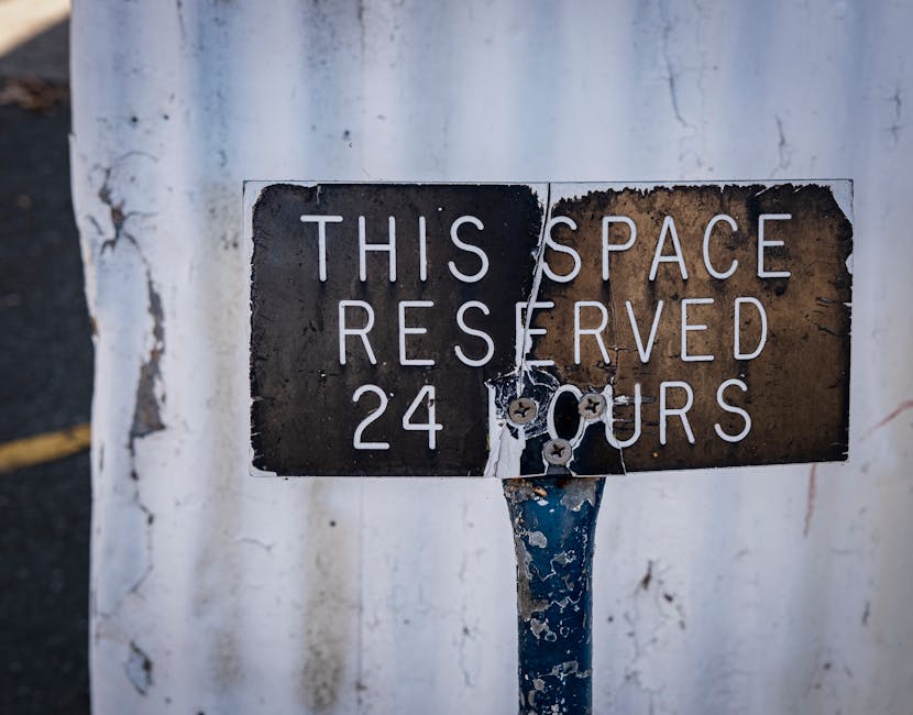 A weathered metal parking permit sign attached to a pole outside a property, indicating a parking reservation for 24 hours. The sign has peeling black and rust-colored paint with white lettering stating 'This Space Reserved 24 Hours'. The background features a rough white wall with visible cracks and chipped paint. This image relates to house removals or relocation services, such as those offered by Man with Van Hornchurch, highlighting the importance of understanding local parking permits and council rules when planning a home relocation or furniture transport within Hornchurch.