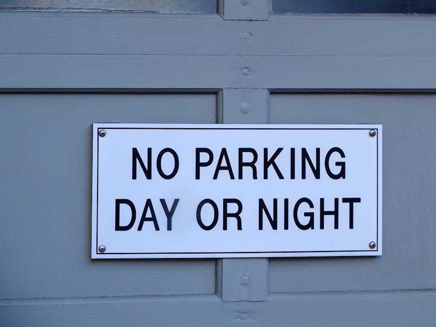 A rectangular white sign with black uppercase text reading 'NO PARKING DAY OR NIGHT' is mounted on a light grey wooden garage door. The sign is secured with four metal screws, one in each corner. The background features the horizontal panels of the garage door, with visible wood grain and subtle panel gaps. The image is well-lit with even natural lighting, emphasizing the sign's clear message and straightforward design. This visual context relates to house removals and moving logistics, highlighting the importance of understanding parking regulations when planning a furniture transport or property relocation, as managed by Man with Van Hornchurch.