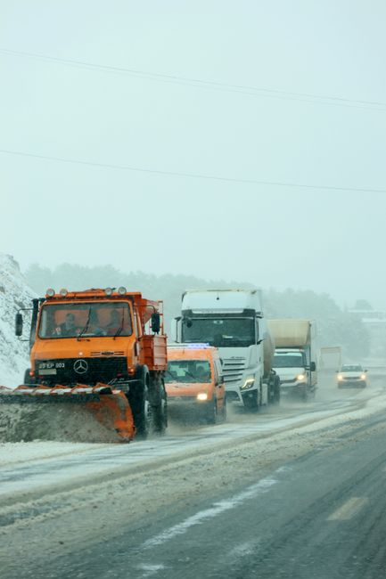 A line of vehicles including a large orange snowplough truck with a wide, angled blade attached at the front, and several other cars and vans, are driving along a snow-covered road under overcast, foggy conditions. The snowplough is actively clearing the road, which is partially covered with snow and slush, with visible snow piles along the edges. The background features a faint outline of trees and power lines, with limited visibility due to the fog. The scene depicts winter weather impacting traffic flow, relevant to the context of house removals and moving logistics, as professional services like Man with Van Hornchurch may be affected by road closures or delays during adverse weather conditions.