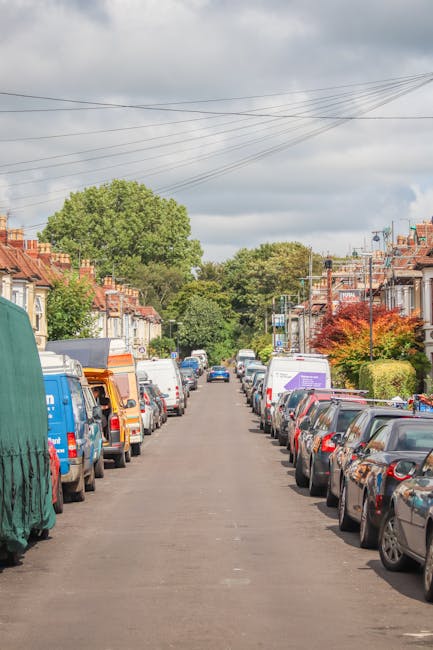 A residential street in Hornchurch lined with rows of parked cars on both sides, including hatchbacks, sedans, and vans, with some vehicles partially covered with protective tarps. The street surface is asphalt, and there are various cardboard boxes and small pieces of packaging materials placed near the vehicle doors, indicating a home relocation process. Residential houses with brick facades, pitched roofs, and small front gardens are visible along the street, with some featuring hedges and trees, including a notable large tree at the end of the street. The sky overhead is cloudy with some patches of sunlight breaking through, and overhead power lines run parallel above the street. This scene reflects a typical setting involved in furniture transport and packing during a house removal. The presence of multiple moving vehicles and the arrangement of the objects suggest an ongoing loading or unloading activity, supporting the context of professional removals and moving services by [COMPANY_NAME], tailored for navigating Hornchurch road closure permit considerations.