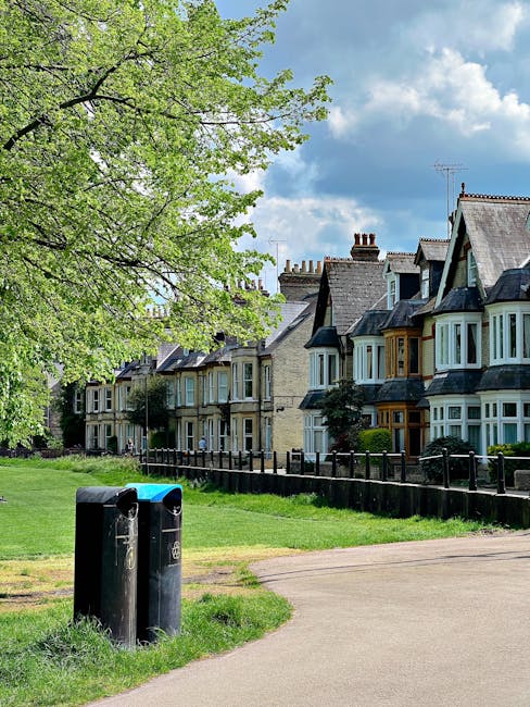 A residential street scene showing a row of terraced houses with characteristic Victorian-style bay windows, some with painted wooden frames, and sloped tiled roofs against a partly cloudy sky. In the foreground, there are two black cylindrical public garbage bins placed on a grassy verge next to a paved footpath. A large leafy green tree with fresh spring foliage extends over the scene from the left side, providing partial shade. The grass appears well-maintained, and a black metal fence runs along the front of the houses, separating the pathway from the gardens. This setting aligns with a typical area where a local house removals service, such as Man with Van Hornchurch, might operate during home relocation or furniture transport activities, potentially involving loading procedures near the curbside as part of a moving process in the Elm Park or Emerson Park area.